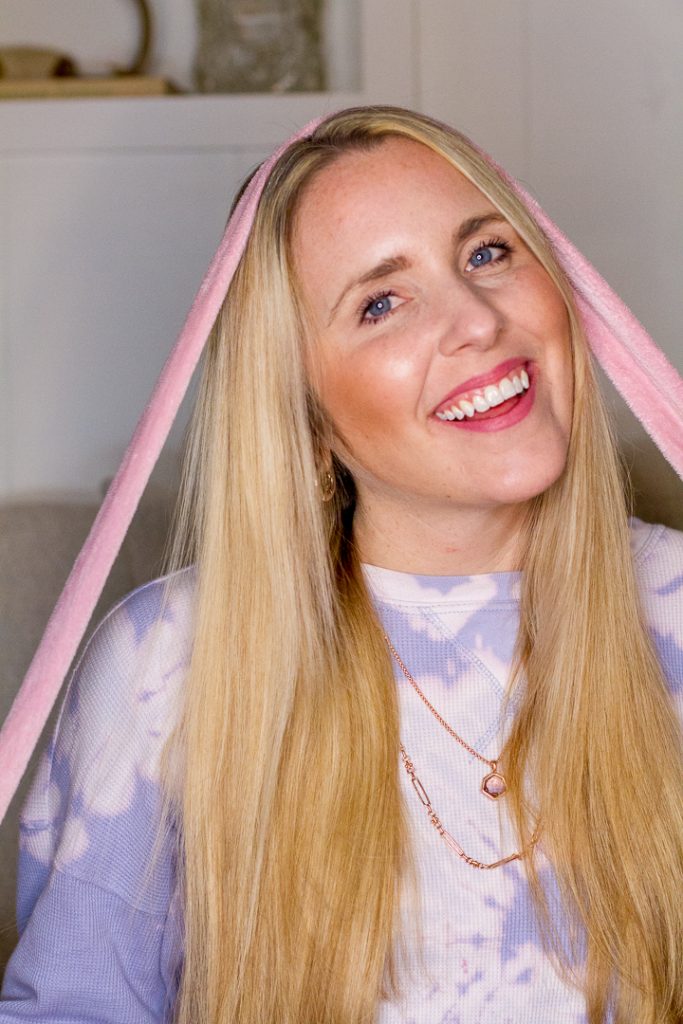 Heatless Waves by popular Nashville beauty blog, Pearl and Twirls: image of a woman with long straight blonde hair and wearing a purple tie dye shirt while holding a pink bathrobe tie on the top of her head. 