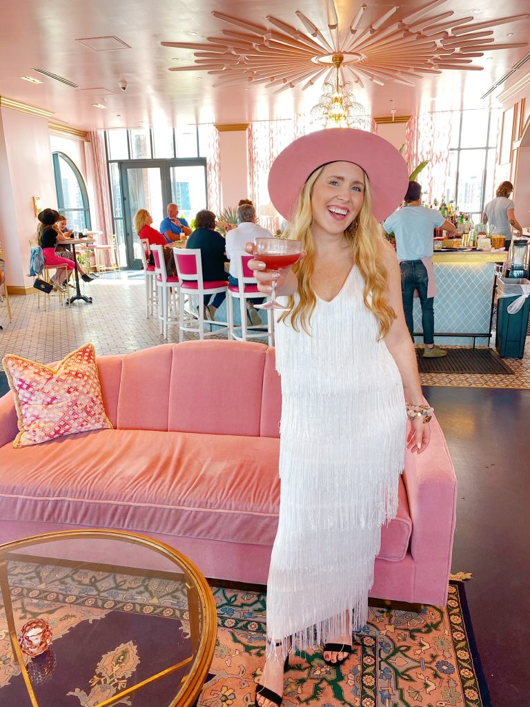 Bachelorette Party Ideas by popular Nashville lifestyle blog, Pearls and Twirls: image of a woman wearing a white finge dress, black heel sandals and a pink felt hat standing in a hotel with a pink velvet couch and pink ceilings and walls. 