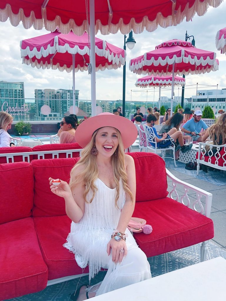 Bachelorette Party Ideas by popular Nashville lifestyle blog, Pearls and Twirls: image of a woman wearing a white fringe dress, pink felt hat and sitting on pink patio furniture under a pink shade umbrella with white fringe. 