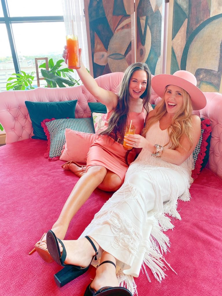 Bachelorette Party Ideas by popular Nashville lifestyle blog, Pearls and Twirls: image of two women sitting on a pink bed and drinking some adult drinks. 