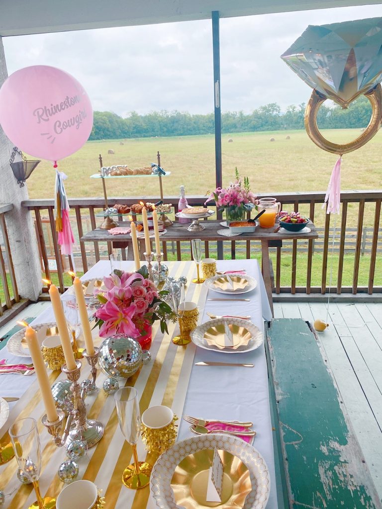 Bachelorette Party Ideas by popular Nashville lifestyle blog, Pearls and Twirls: image of a bachelorette party tablescape, with gold dishes, white and gold stripe table runner, engagement ring balloon, gold and white dishes, and a pink floral arrangement. 