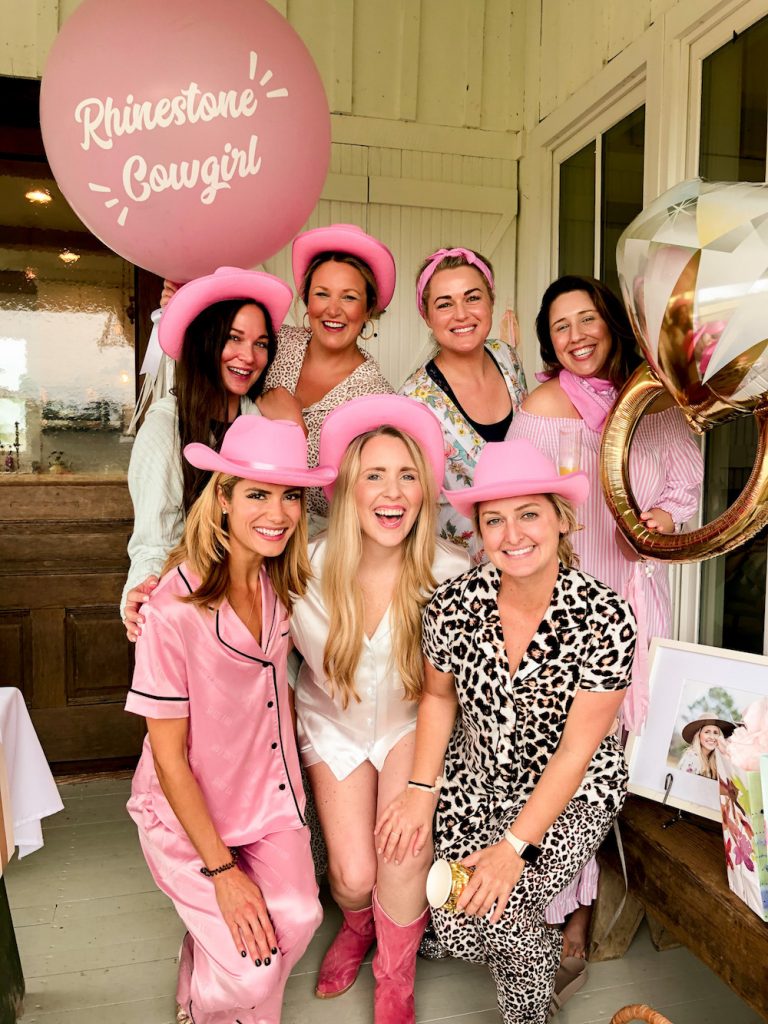 Bachelorette Party Ideas by popular Nashville lifestyle blog, Pearls and Twirls: image of a group of women wearing pajamas, pink cowgirl hats and holding a Rhinestone Cowgirl helium ballon, and engagement ring helium balloon. 