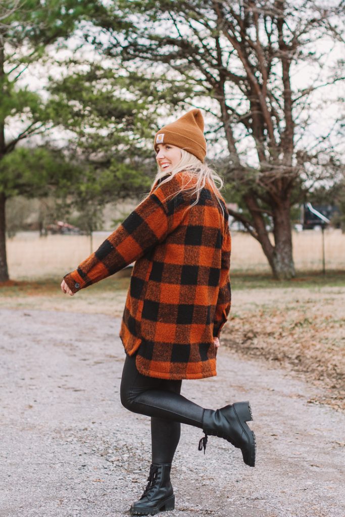 shacket outfit with boots |shacket and leggings outfit | Old Navy Shacket by popular Nashville fashion blog, Pearls and Twirls: image of a woman standing outside on a gravel road and wearing a Carhart beaning, Old Navy orange and black buffalo plaid shacket, black Spanx faux leather leggings, Target combat boots, and My Kind of Lovely swing top.